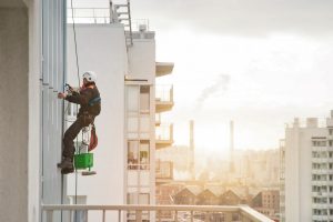 Industrial mountaineering worker in uniform hangs over residential facade building, washing exterior glazing